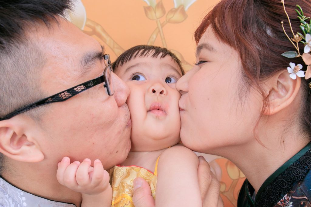 媽媽和爸爸親吻寶寶臉頰，寶寶露出可愛逗趣的表情，溫馨又幽默的家庭攝影瞬間